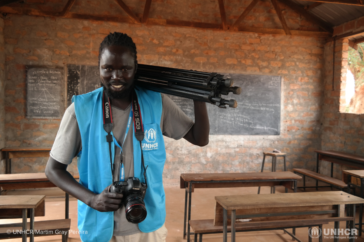 South Sudanese refugee Achouth standing in a refugee camp class room holding his photography equipment